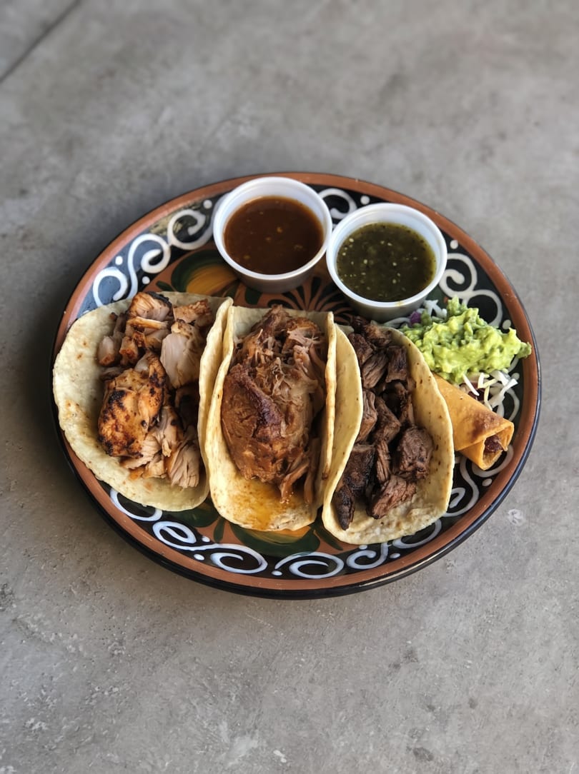 Three tacos with different meats, guacamole, and two sauces on a decorative plate.